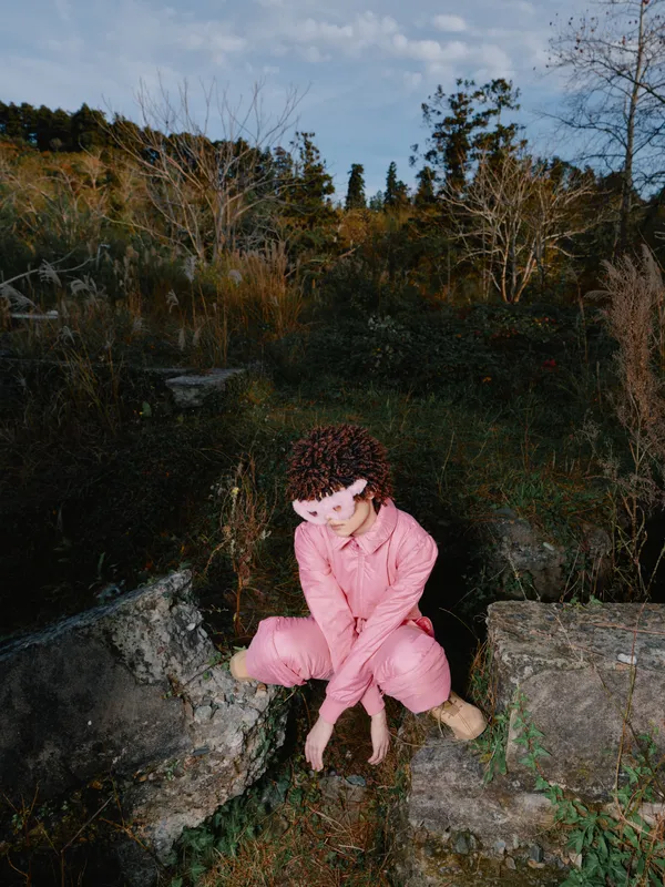 Outdoor nature scene with a girl sitting on rocks in a pink jumpsuit, calm expression, earthy tones surrounding trees and bushes, serene portrait outdoors