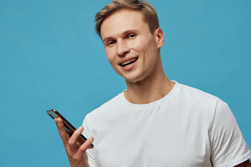 smiling young caucasian man hold smartphone use voice assistant gesture blue background casual white t-shirt technology concept