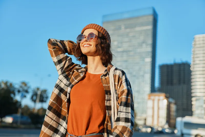 Woman with smile in urban scene wearing sunglasses and beanie and plaid jacket, joyful candid moment in golden hour glow, authenticity and mindful living captured in emotional storytelling.