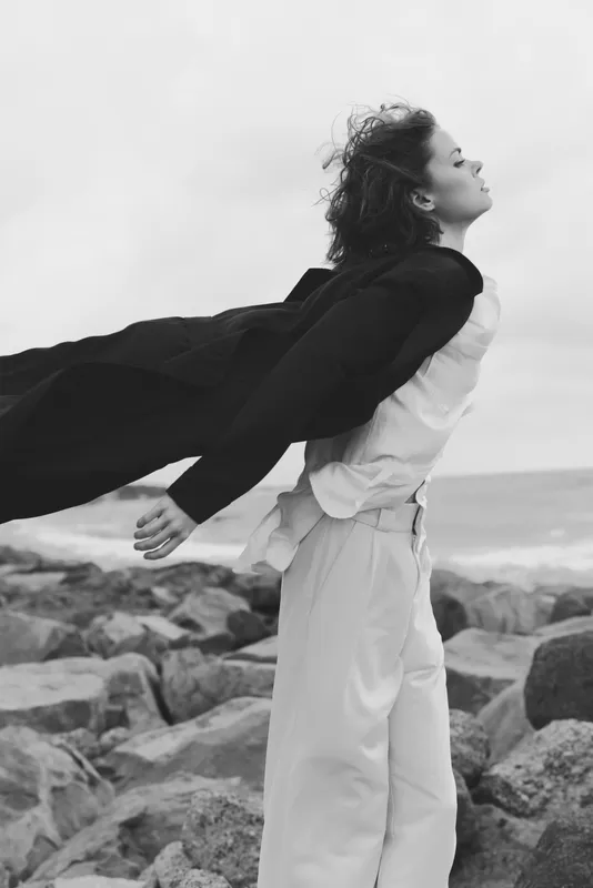 Wind catches an open coat as a woman stands on rugged rocks by the sea, embracing freedom and movement in a moody black and white moment.
