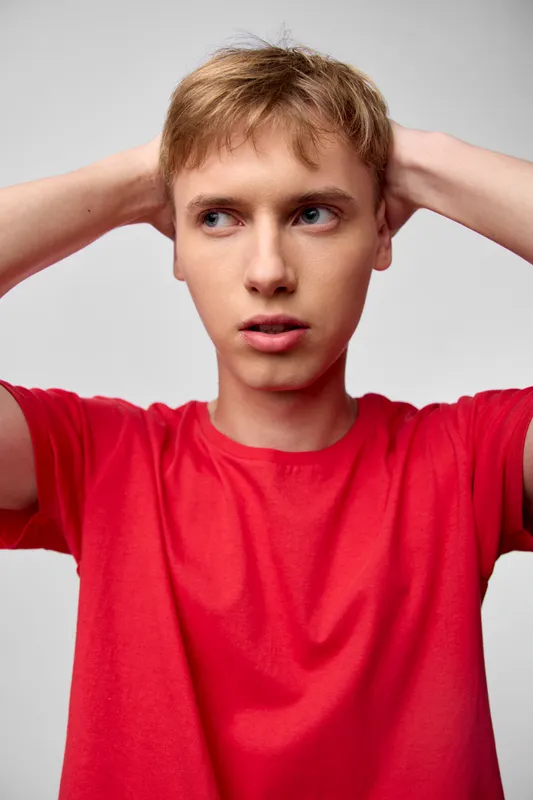 Thoughtful, tense moment of a young man with hands on his head, wearing a red shirt in a studio setting, conveying confusion, stress, and contemplation through facial cues, body language, and
