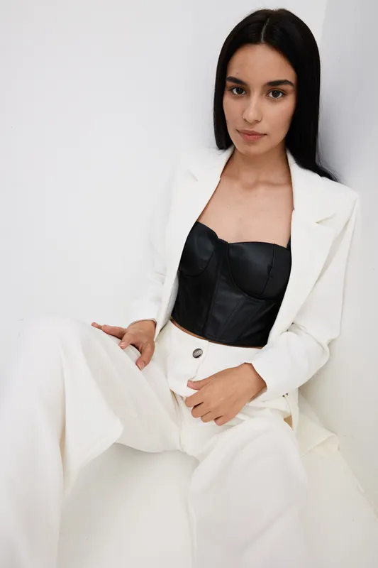 Young woman in stylish black corset and white pantsuit sits relaxed against white wall, showcasing modern fashion with natural makeup and straight dark hair, minimalistic background.
