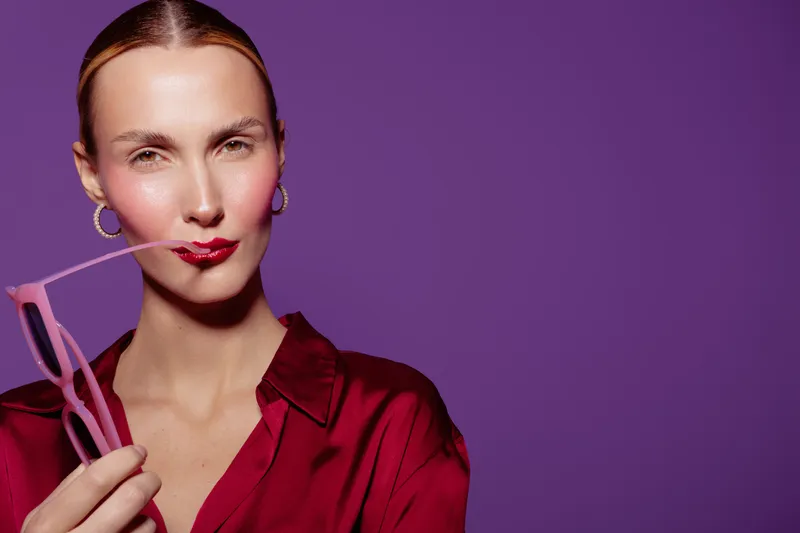 Confident woman in red blouse holds pink glasses near lips, posing against purple background. Concept of fashion, style, beauty, and modern portrait photography with vibrant colors.