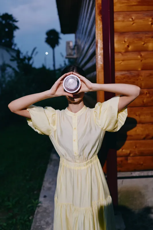 Young woman in a pale yellow dress holds a camera lens to her face outdoors near a wooden cabin at dusk, creating a playful mysterious portrait with moody light