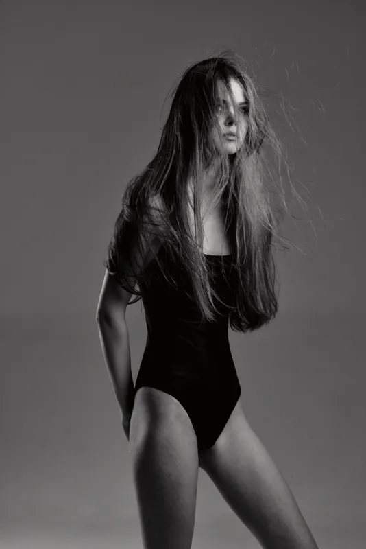 Black and white portrait of a tall woman with long hair wearing a black bodysuit and expressive pose in studio lighting.
