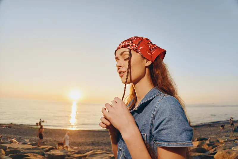 Lifestyle woman in modern boho-western denim street style with red bandana, enjoying calm sunset by the beach, cinematic warm film color tones enhancing mood.