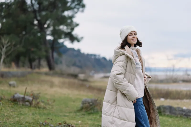 Winter coat, woman in a scarf and beanie walking in an open park, calm landscape with distant trees, soft natural light, cozy fashion moment outdoors, serene smile and breeze