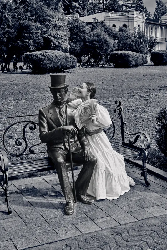A vintage couple sits on a park bench, the man wearing a top hat and suit, the woman in a flowing dress with a fan, creating a romantic nostalgic outdoor moment.