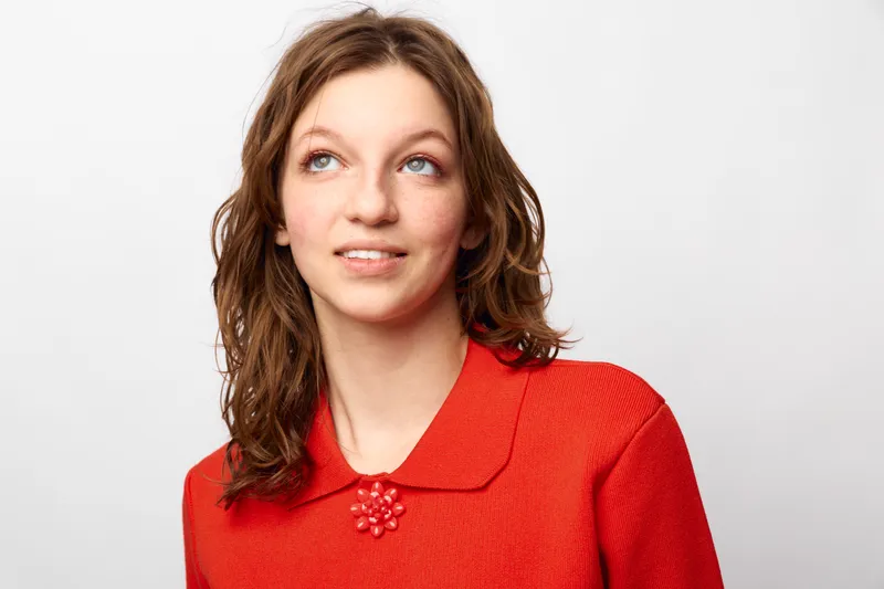 Young woman with wavy brown hair wearing a red sweater with a decorative brooch looks up and smiles slightly against a plain white background. Concept of casual style and optimism.
