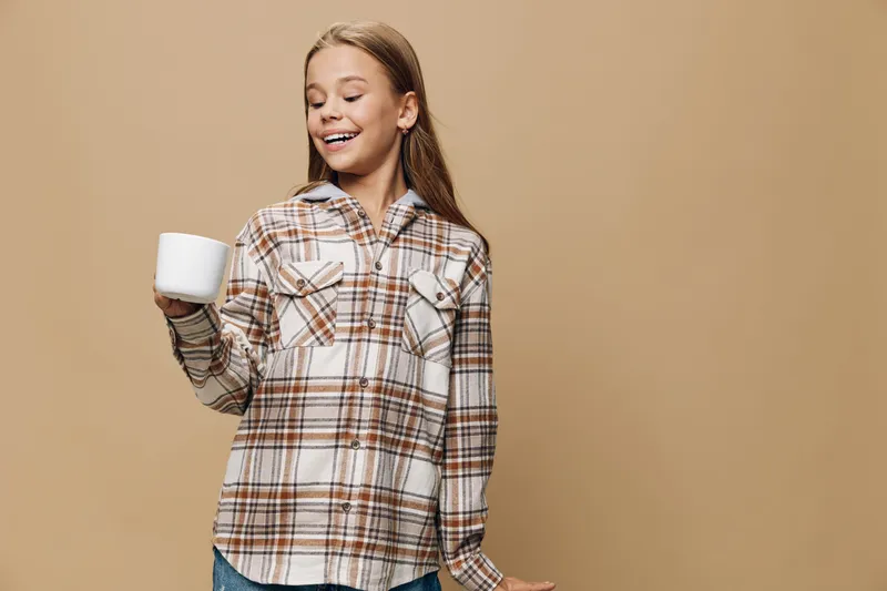 Girl with long hair smiling and holding white cup wearing plaid shirt standing against beige background with casual style and happy expression.