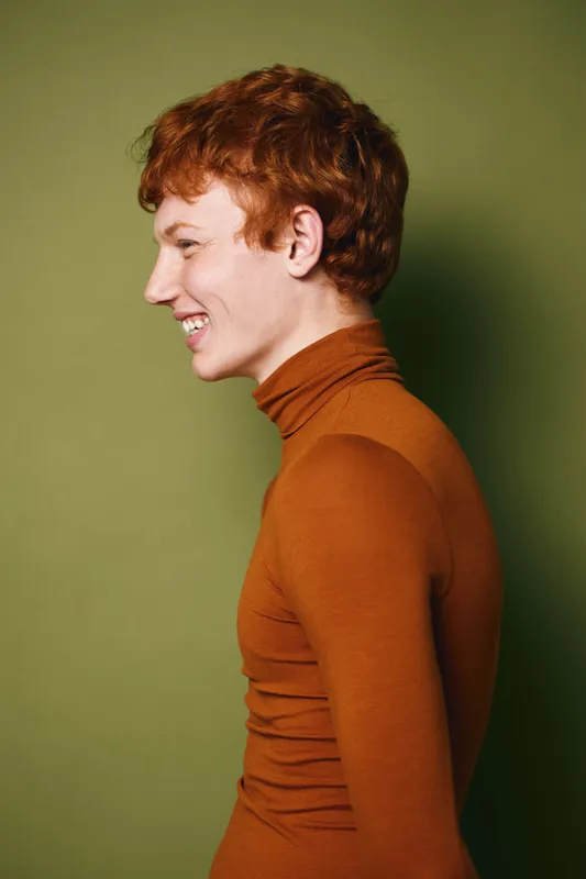 Young man with red hair wearing orange turtleneck smiling sideways on green background with casual hairstyle in studio portrait, happy and relaxed expression.