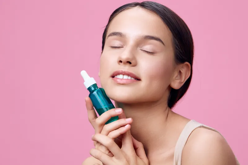 Young woman with a skincare serum, expressing joy against a pink background, highlighting beauty and wellness in the skincare concept