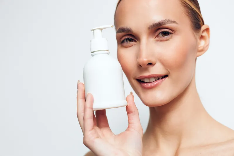 Young woman holding a white skincare bottle by her face, serum for hydration, gentle beauty smile, cosmetic care, clean white background, soft daylight, minimal