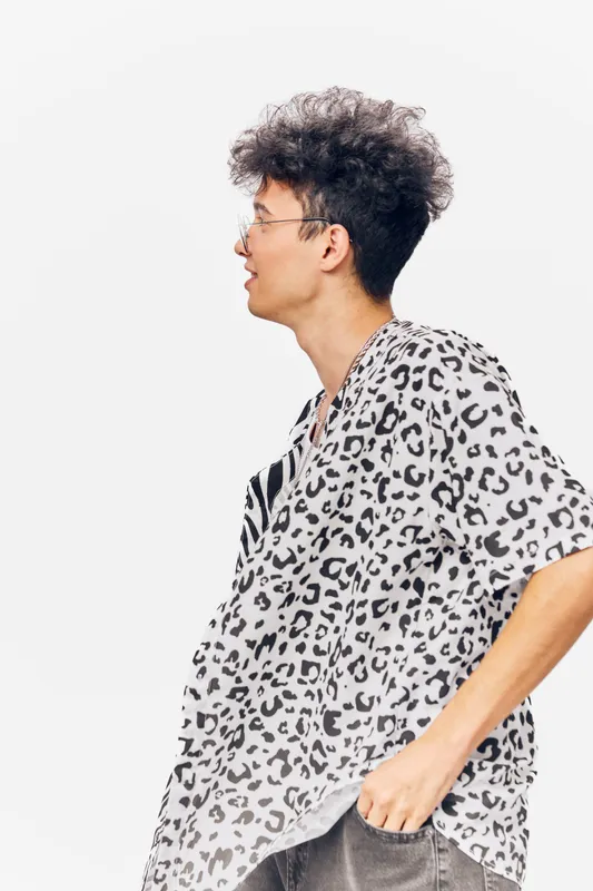 Young man with curly hair wearing leopard print shirt and glasses, standing sideways against white background, casual style and thoughtful expression.