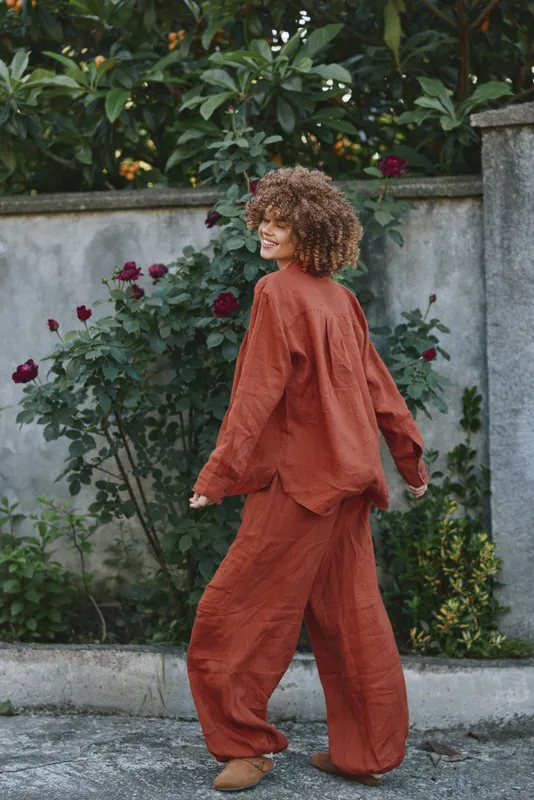 Curly-haired woman in a lounge orange set walking outdoors among garden greenery, casual lifestyle moment, natural light, relaxed, cheerful mood, street vibe