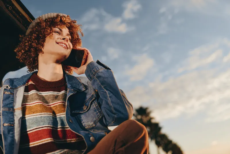 Woman lifestyle outdoors in sunlight, lowangle portrait of smiling young, curly auburn hair, denim jacket and sweater, drinking bottle, summer breeze, blue sky clouds, soft bokeh