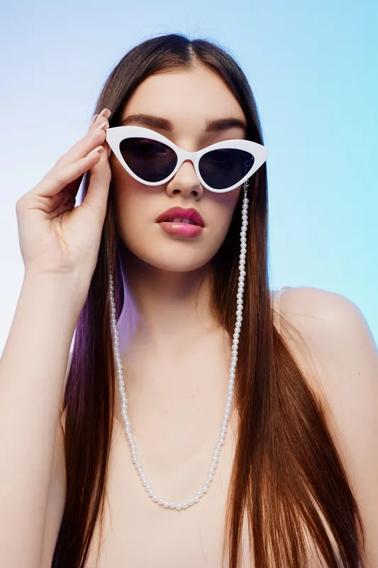 Young woman with long brown hair wearing white sunglasses and a pearl chain poses against a pastel blue background. Concept of beauty, fashion, and modern style.