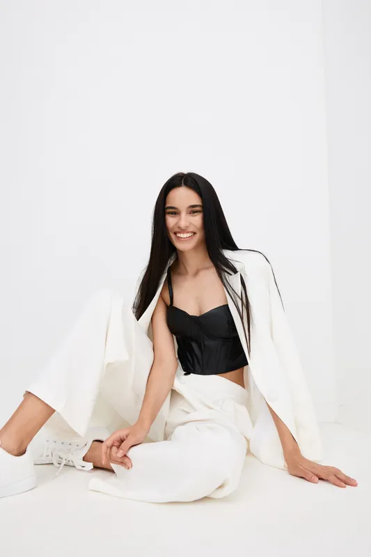Smiling woman in white pants and black top sitting on the floor against a white background, showcasing modern fashion and casual style with natural long dark hair.