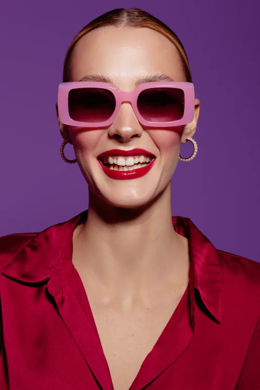 Smiling woman with pink sunglasses and red lipstick wears red satin blouse against purple background. Fashion portrait with bright colors and confident expression. Concept of style and beauty.