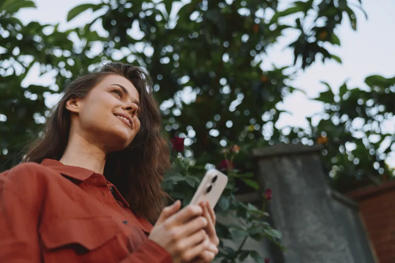 Cheerful woman outdoors, holding smartphone, casual shirt, sunlit garden ambiance, relaxed pose, candid moment, natural colors, lush greenery background, peaceful mood
