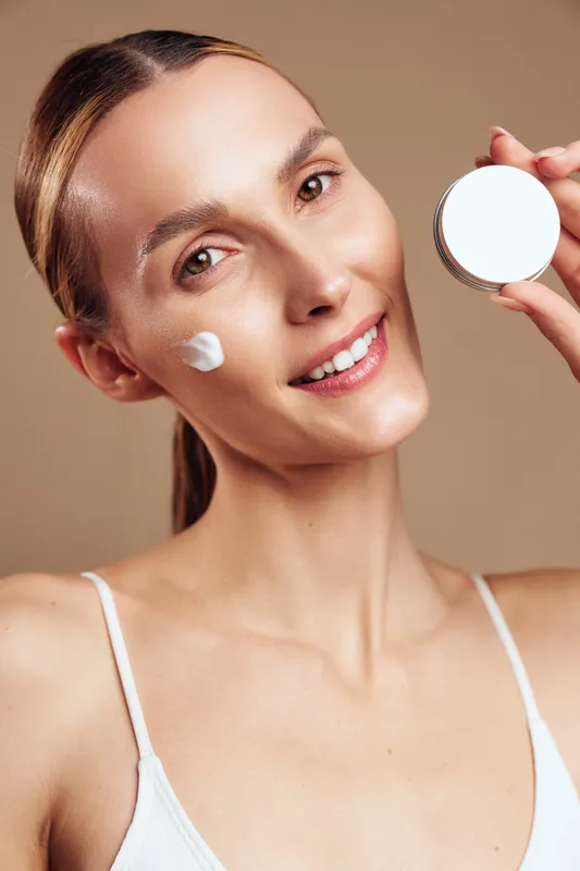 Young woman applying cream on face smiling looking at camera natural skincare routine healthy skin soft lighting beige background beauty concept