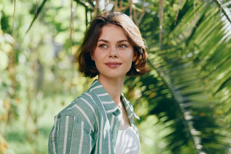 Woman, portrait, outdoor setting, cheerful smile, relaxed pose, natural light, green foliage, striped shirt, short hair, calm mood, bright atmosphere, hopeful vibe, ready for a journey