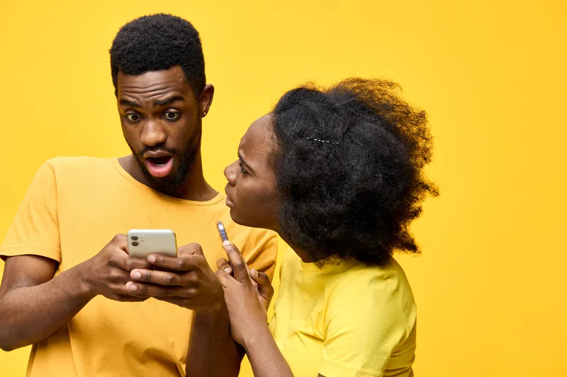 Excited young couple with mobile phones on a bright yellow background, expressing surprise and curiosity while interacting socially