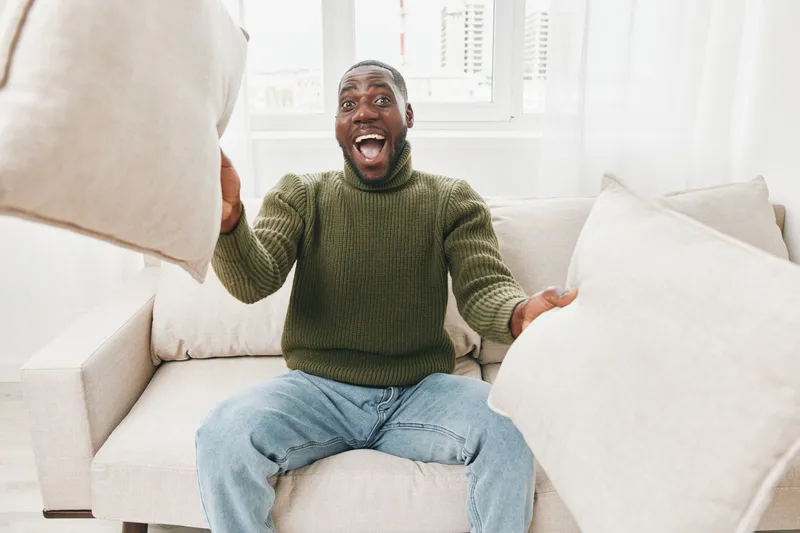 Joyful man in a green sweater playfully tossing cushions on a white sofa, creating a lively and cheerful atmosphere in a bright living room
