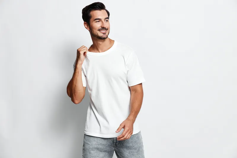 Smiling man in casual white t-shirt and jeans stands confidently while holding his shirt. Bright cheerful atmosphere, isolated on plain light background, studio portrait.