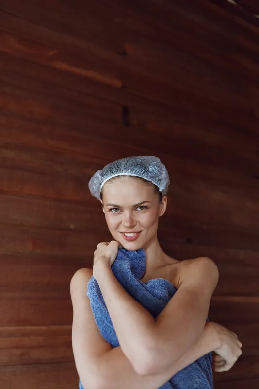 smiling woman wrapped in blue towel with hair net standing against wooden wall, relaxed and fresh after spa treatment, natural skin and calm expression