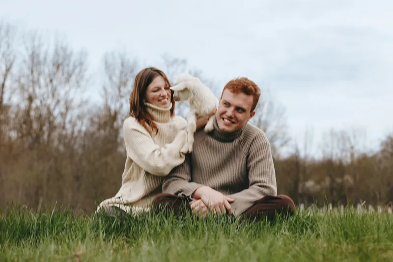 A couple sits on a grassy meadow with a playful goat kid perched on the man’s shoulder, both smiling. A cozy, candid outdoor moment illustrating affection, companionship, and simple rural lifestyle.