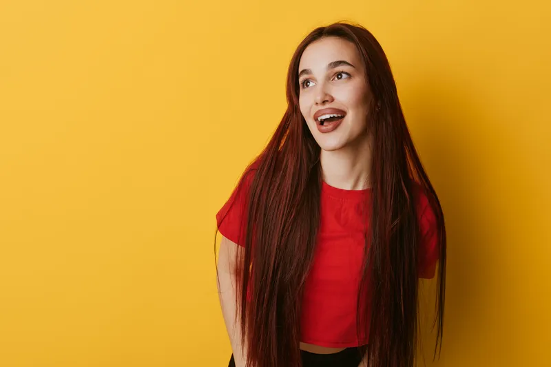 Young woman with long hair wearing red shirt smiling and looking away on yellow background. Portrait of happy female with casual style and natural expression in studio.