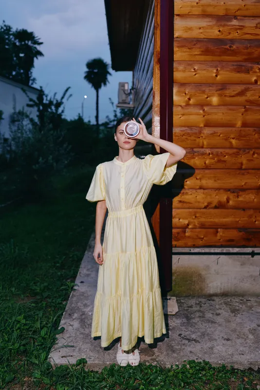 Woman in a pale yellow dress posing outdoors beside a wooden building at dusk, covering one eye with a round prop, creating a mysterious fashion moment.