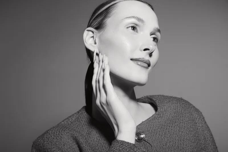Portrait of a fair skinned Caucasian woman with slicked back hair posing thoughtfully with hand near face. Black and white studio shot with soft lighting and elegant mood.