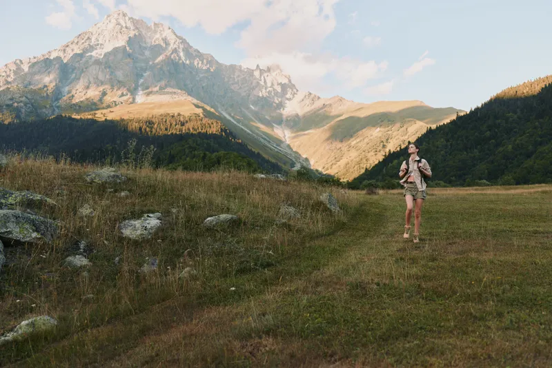 Woman hiker stands in a grassy valley with rugged mountains in the background, wearing hiking gear. A lifestyle moment of outdoor exploration and adventure.