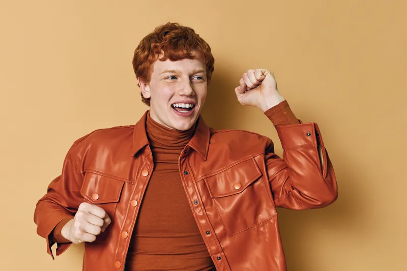 Young man with red hair wearing orange jacket and turtleneck dancing with joy and excitement against beige background. Happiness, celebration and positive energy indoors.