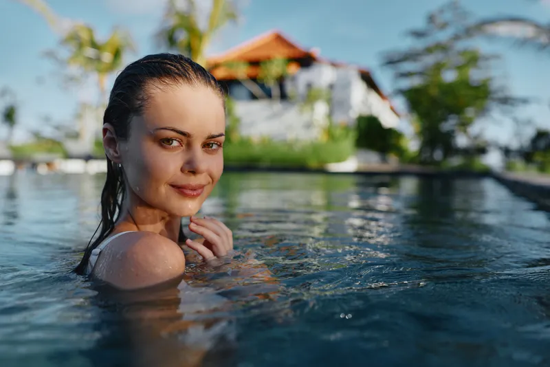 Outdoor pool portrait of a smiling woman enjoying sunny vacation vibes, emerging from clear water relaxed and carefree with a tropical resort backdrop for a lively vacation