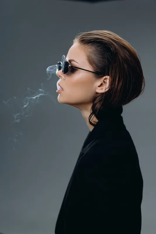 Profile of a stylish woman wearing sunglasses exhaling smoke against a gray background in a dark outfit.
