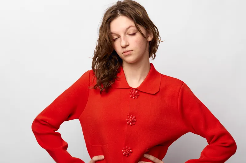 Young woman in red sweater looks down with closed eyes and hands on hips. Studio portrait on white background. Concept of contemplation, mood, and casual fashion.