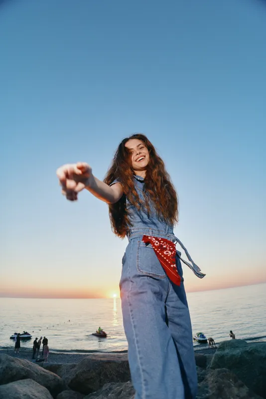 smiling woman in denim jumpsuit with red bandana enjoying sunset at beach with calm sea and clear sky, joyful and relaxed mood in nature