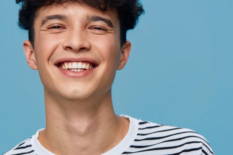 smiling man with striped shirt expressing happiness and joy against plain blue background, casual young adult portrait with natural lighting and positive mood