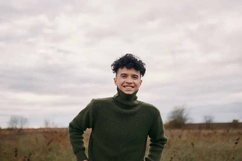 Young man in a green turtleneck smiles confidently in an open field, a candid portrait emphasizing natural expression, authenticity and credibility in an outdoor setting.