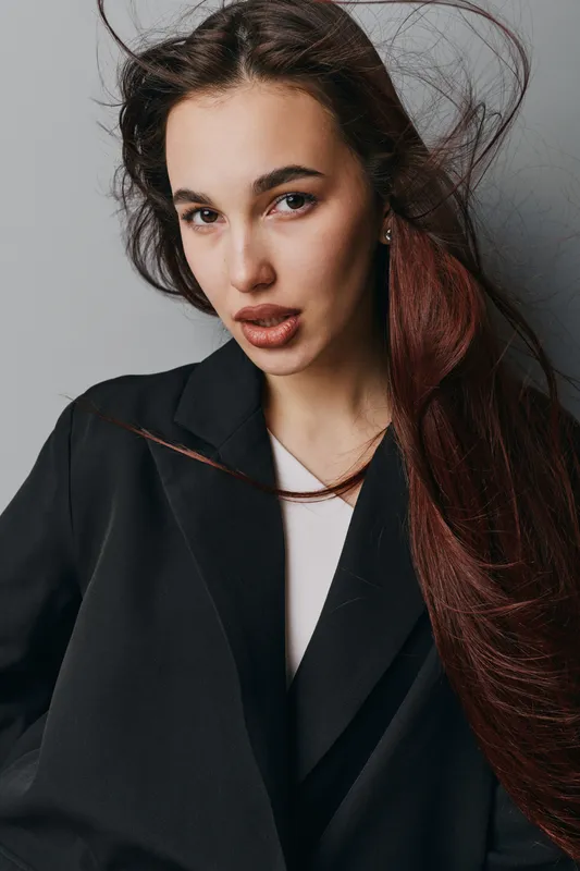 Woman with long brown hair and natural makeup wearing black coat posing indoors. Portrait of young female model with serious expression and loose hair against gray background.