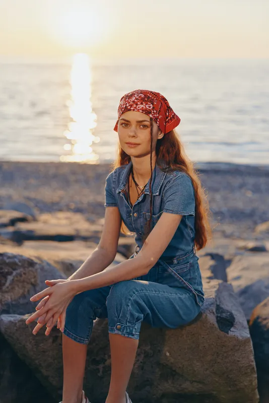 Lifestyle woman in modern boho-western denim street style with red bandana sitting on rocks by the sea at sunset, film color warm tones, natural light, relaxed mood.