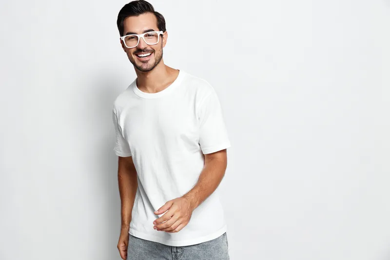 Smiling man wearing a white t-shirt stands against a plain solid background, looking at the camera with a joyful expression, conveying a sense of happiness and approachability.