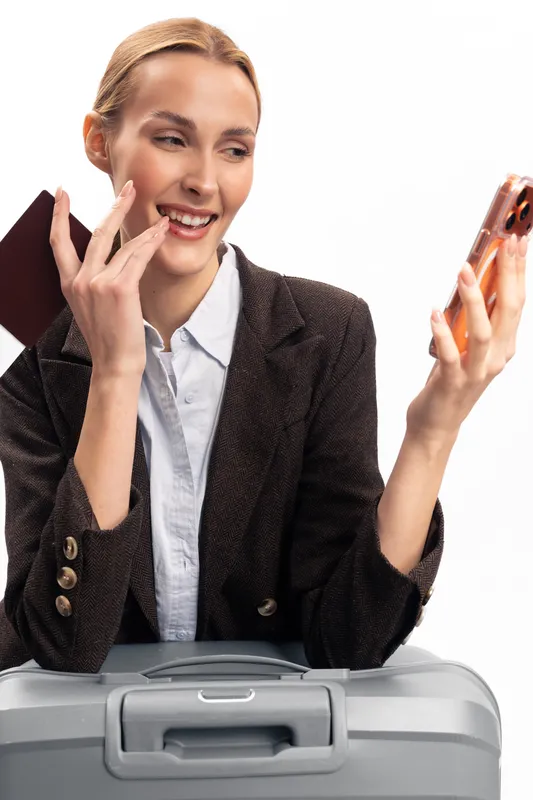 Smiling woman with suitcase holding passport and smartphone, making a video call. Concept of travel, communication, and business trip preparation in modern lifestyle.