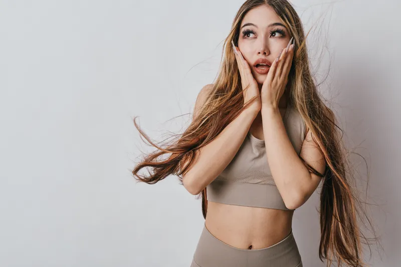 Young woman with long hair and sporty outfit posing with hands on cheeks expressing surprise on gray background in studio. Female model looking up with open mouth and emotion.