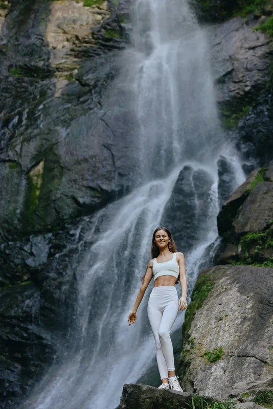 young woman standing near a large waterfall in white activewear, surrounded by rocks and lush green nature, embodying fitness and adventure outdoors.