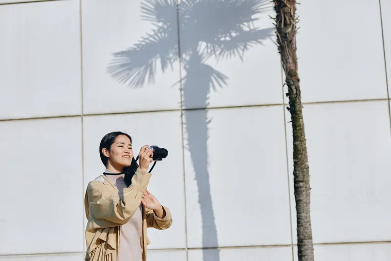 Photographer, woman with camera gazing upward in a bright outdoor setting beside a tall palm shadow against a modern wall, while clear daylight highlights textures and color.