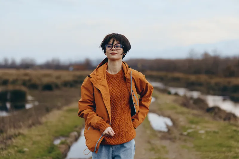 Woman lifestyle outdoors, standing on riverbank path, wearing orange jacket and glasses, relaxed smile, hands in pockets, soft daylight, natural landscape, calm mood, fresh breeze